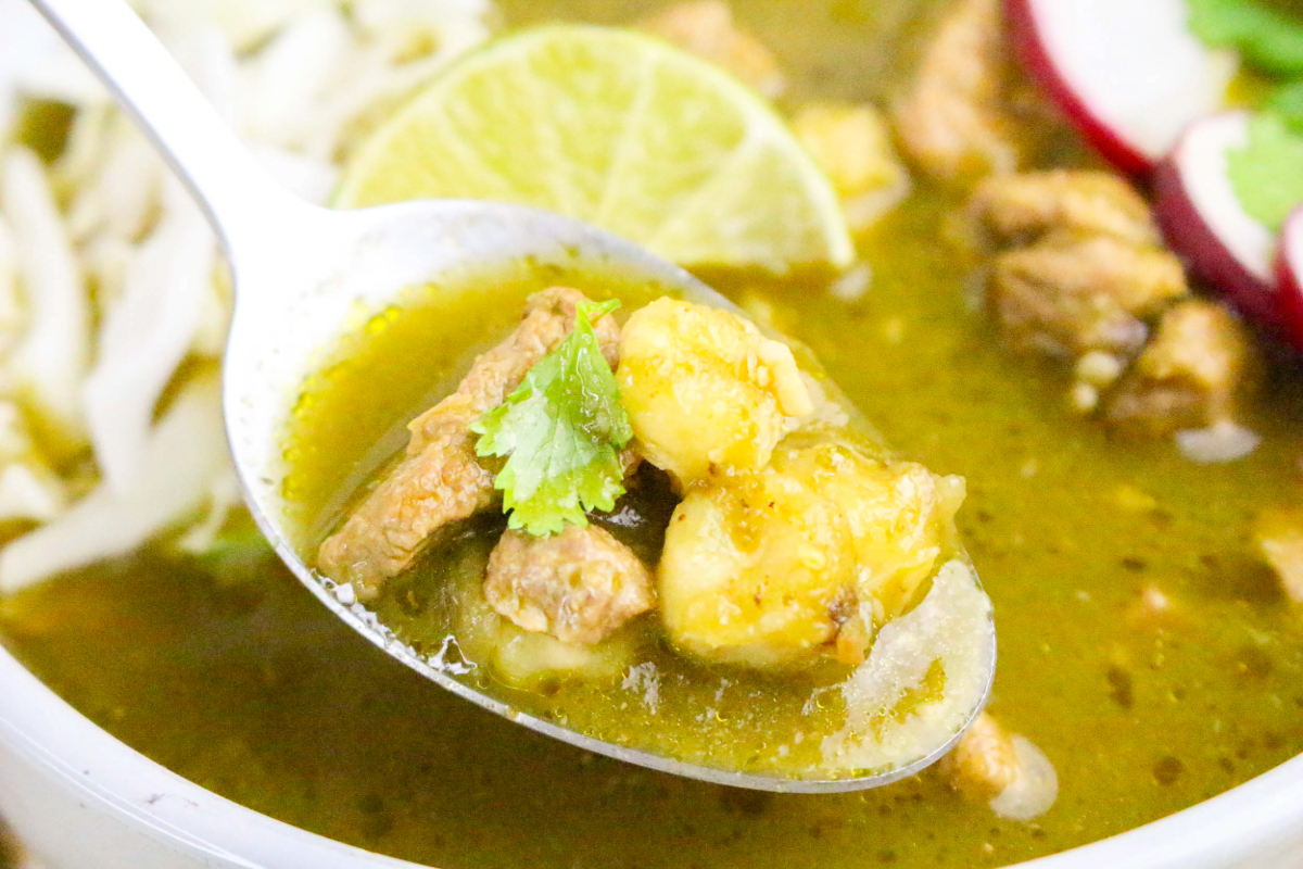 A stainless spoonful of hominy and pork from a white soup bowl.