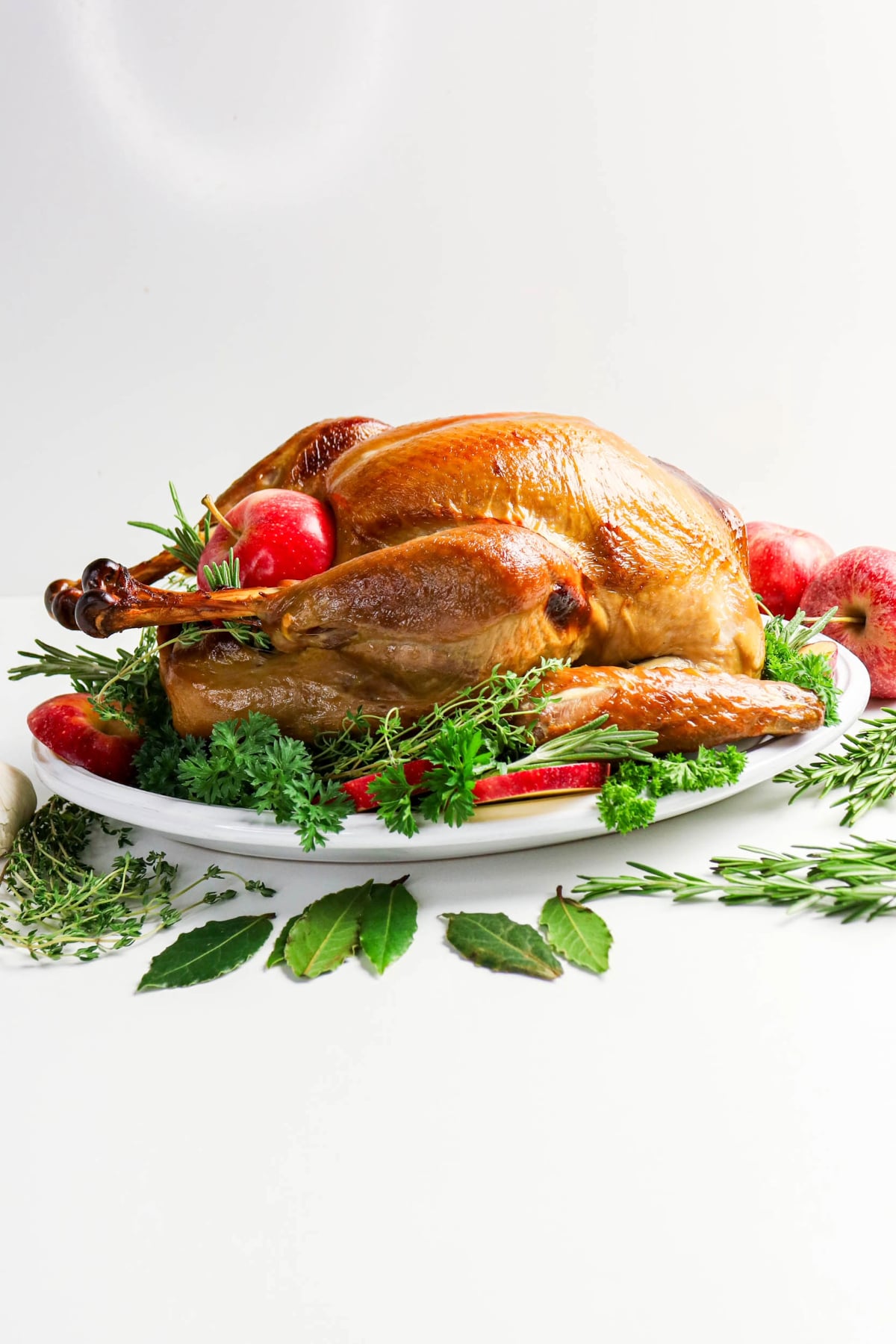 a whole cooked turkey on a white platter with fresh herbs and sliced apples.