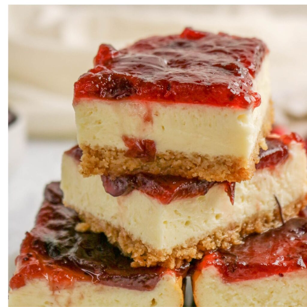 Three cheesecake bars with a graham cracker crust are stacked, each topped with a glossy layer of red fruit jam. The edges show creamy cheesecake filling and crumbly crust layers.