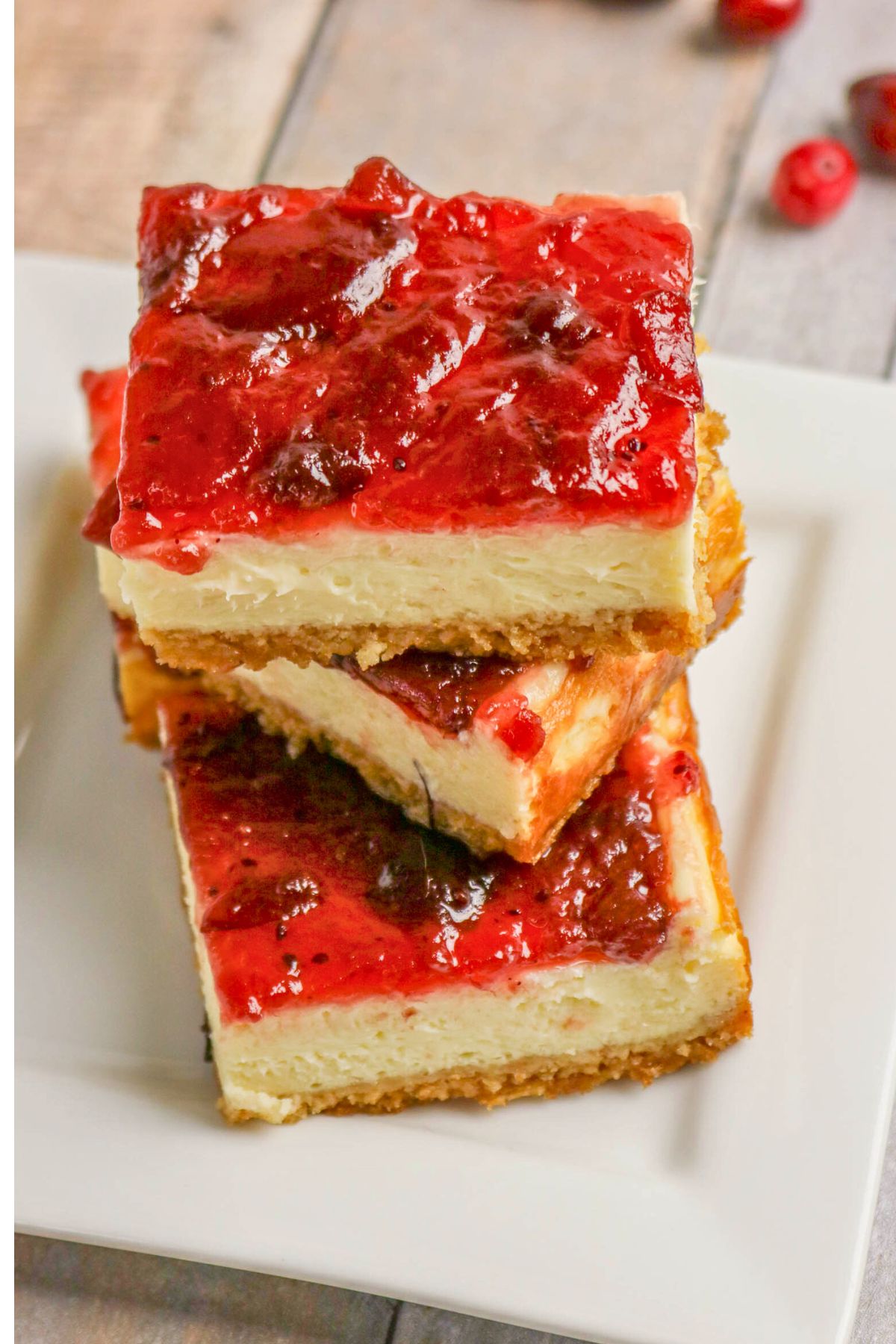 Three cheesecake bars with a crumbly graham cracker crust, creamy filling, and a layer of red fruit topping are stacked on a white plate.