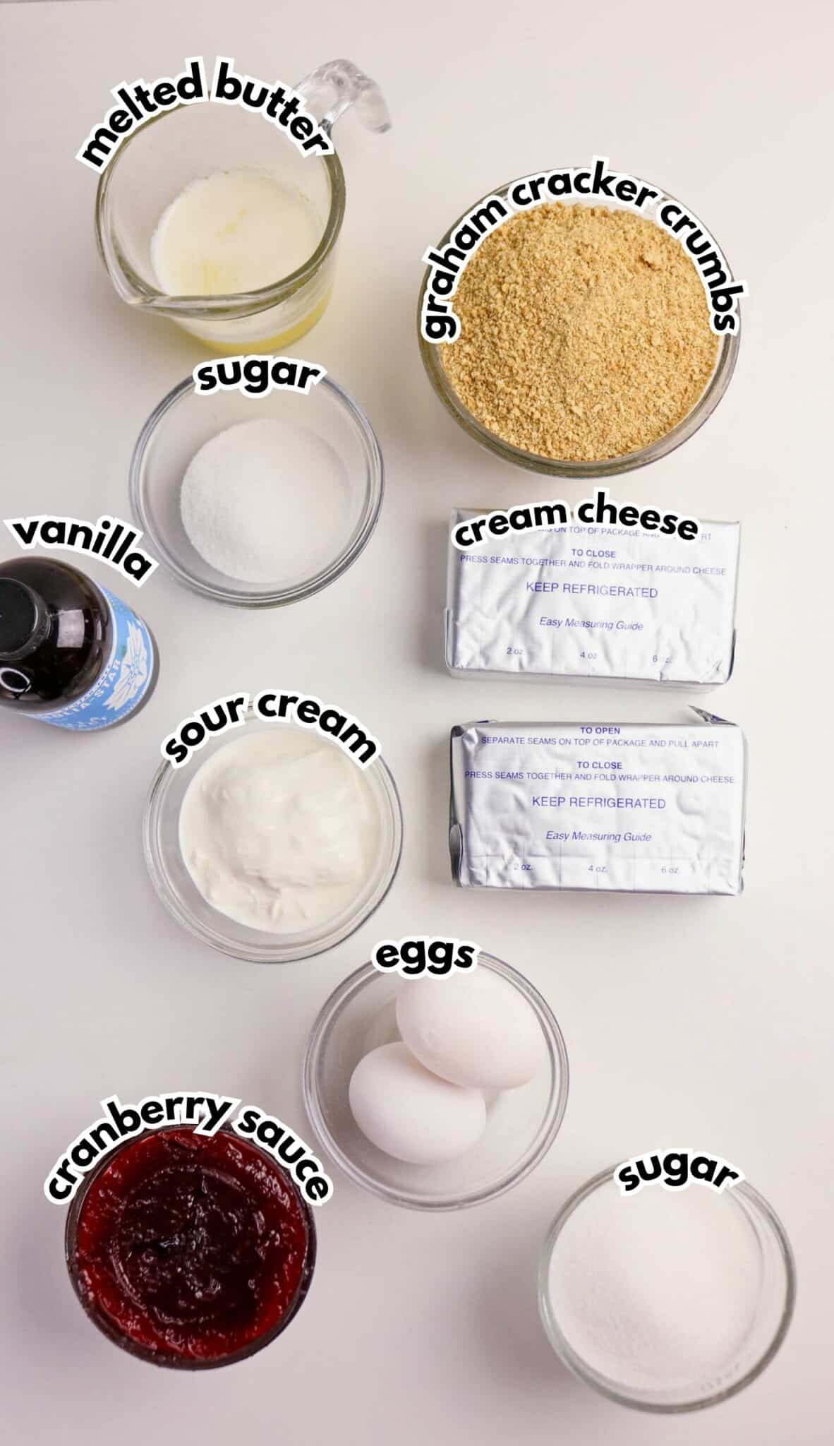 Baking ingredients on a white surface, including melted butter, graham cracker crumbs, two blocks of cream cheese, vanilla, sour cream, cranberry sauce, three eggs, and two bowls of sugar.