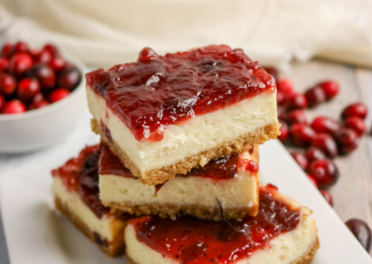 Three cheesecake bars with a graham cracker crust and a layer of cranberry sauce on top are stacked on a white plate, with fresh cranberries in the background.