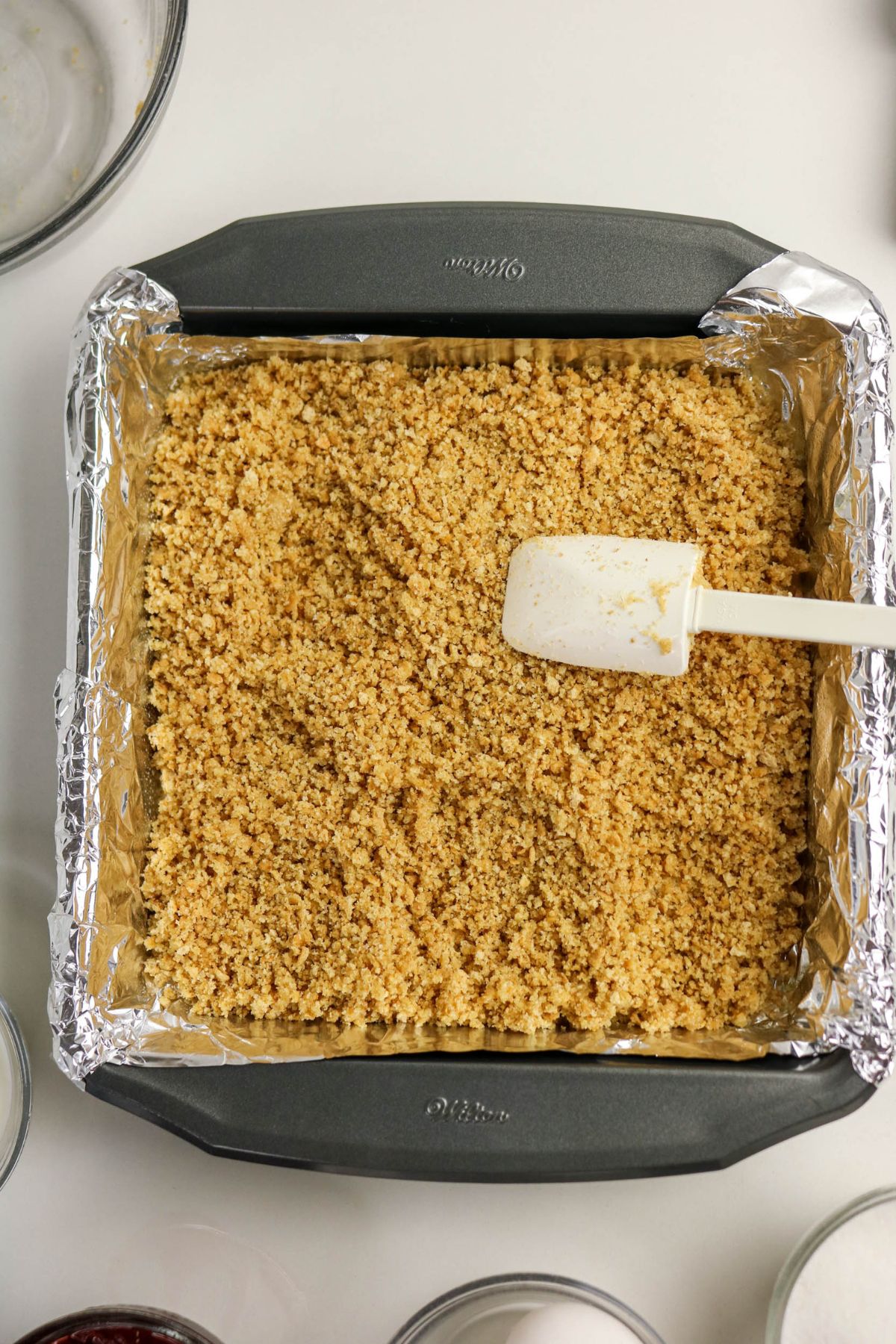 A square baking pan lined with aluminum foil and filled with an even layer of crumbly mixture, being spread with a white spatula.