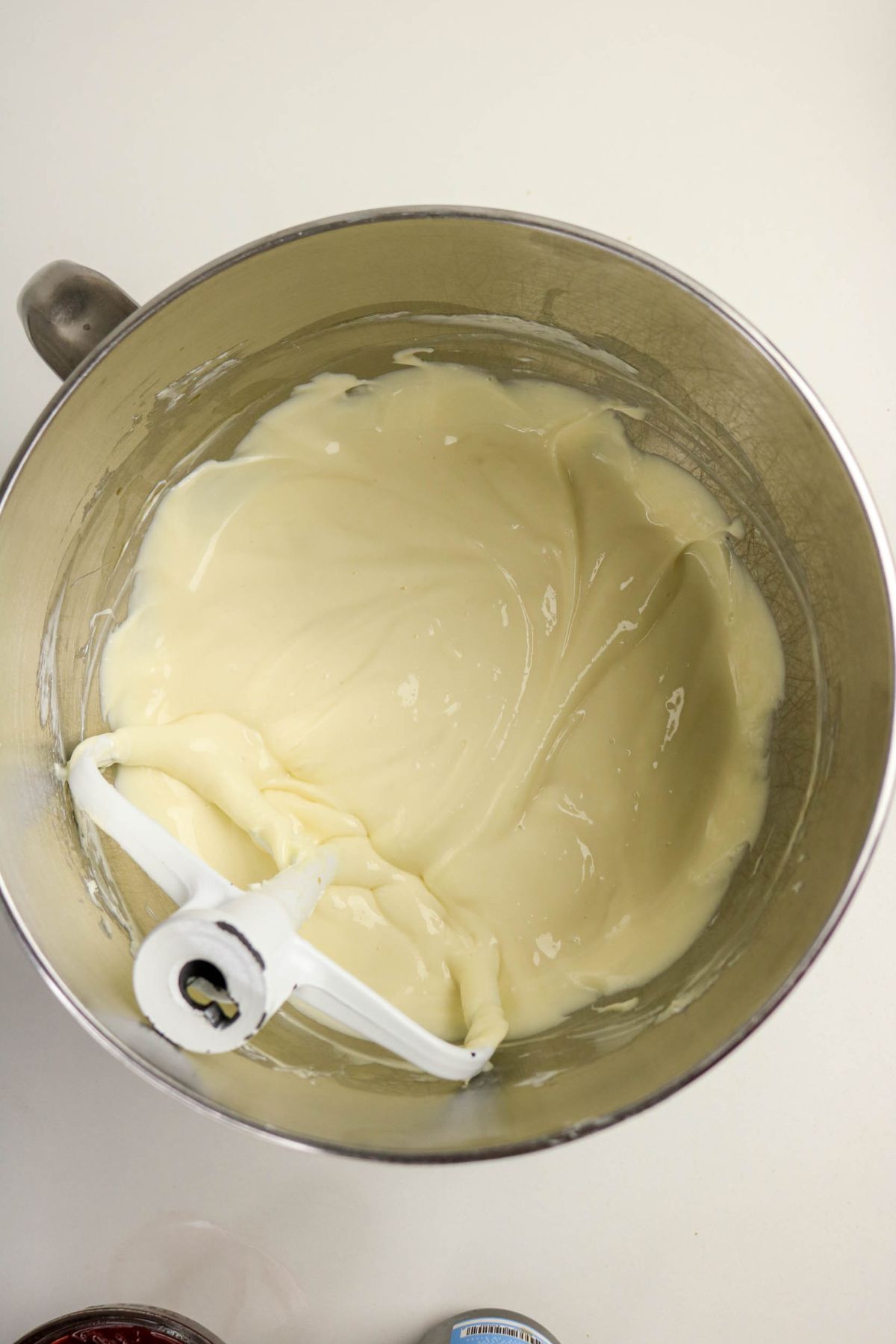 A metal mixing bowl with creamy, cheesecake batter and a white paddle attachment resting inside, set on a white countertop.