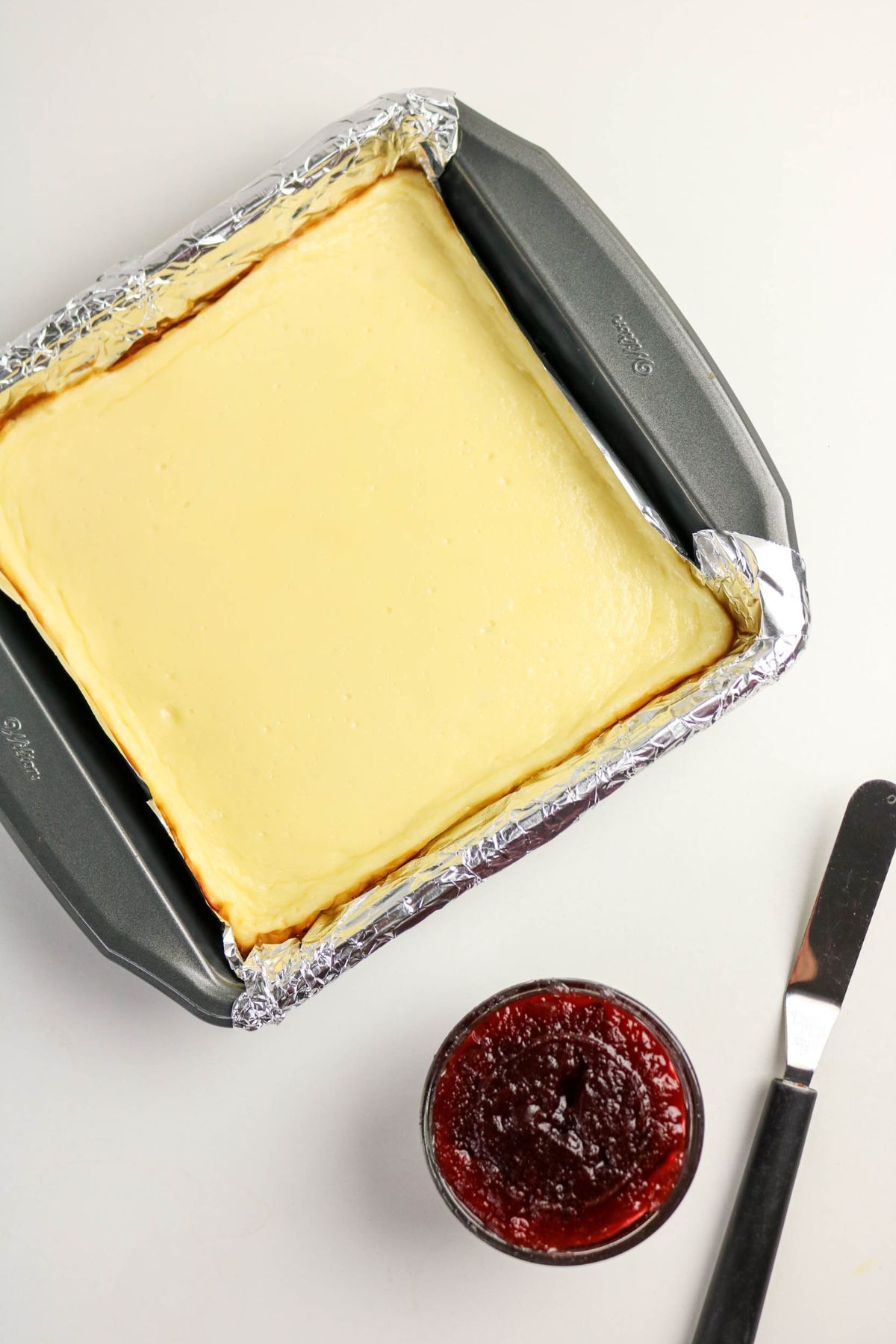 A square cheesecake baked in a foil-lined pan sits on a white surface next to a small glass bowl of cranberry sauce and a butter knife.