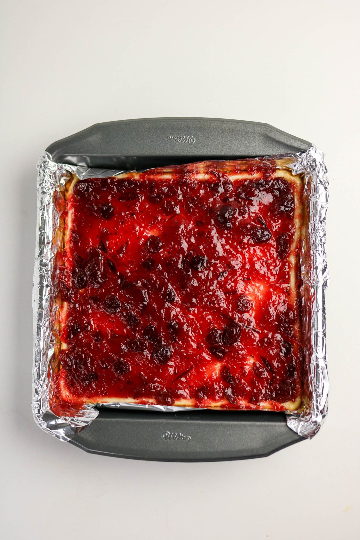 Cranberry sauce being spread on top of bars in a foil-lined baking pan.
