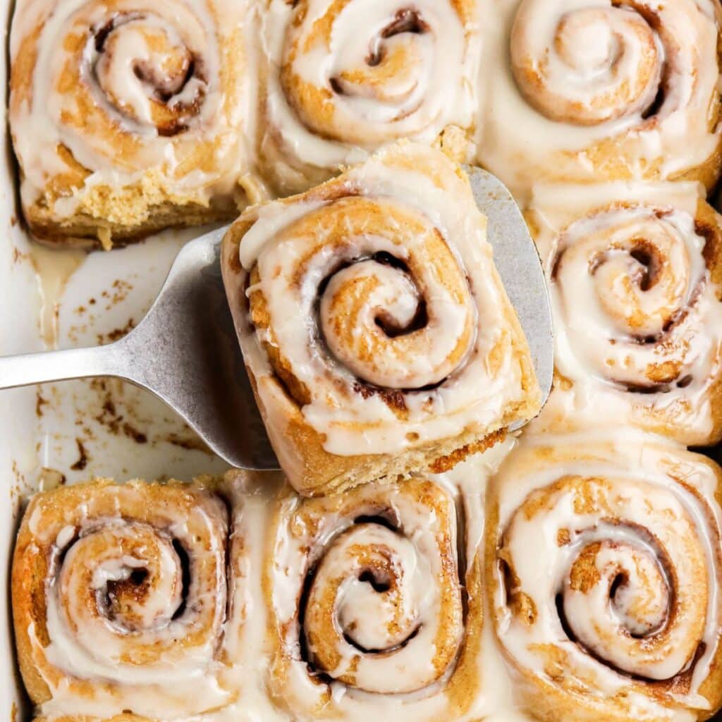A pan of iced cinnamon rolls, with one cinnamon roll being lifted out on a metal spatula. The rolls are golden brown and covered with a layer of white icing.