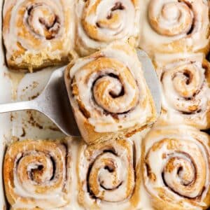 A pan of iced cinnamon rolls, with one cinnamon roll being lifted out on a metal spatula. The rolls are golden brown and covered with a layer of white icing.