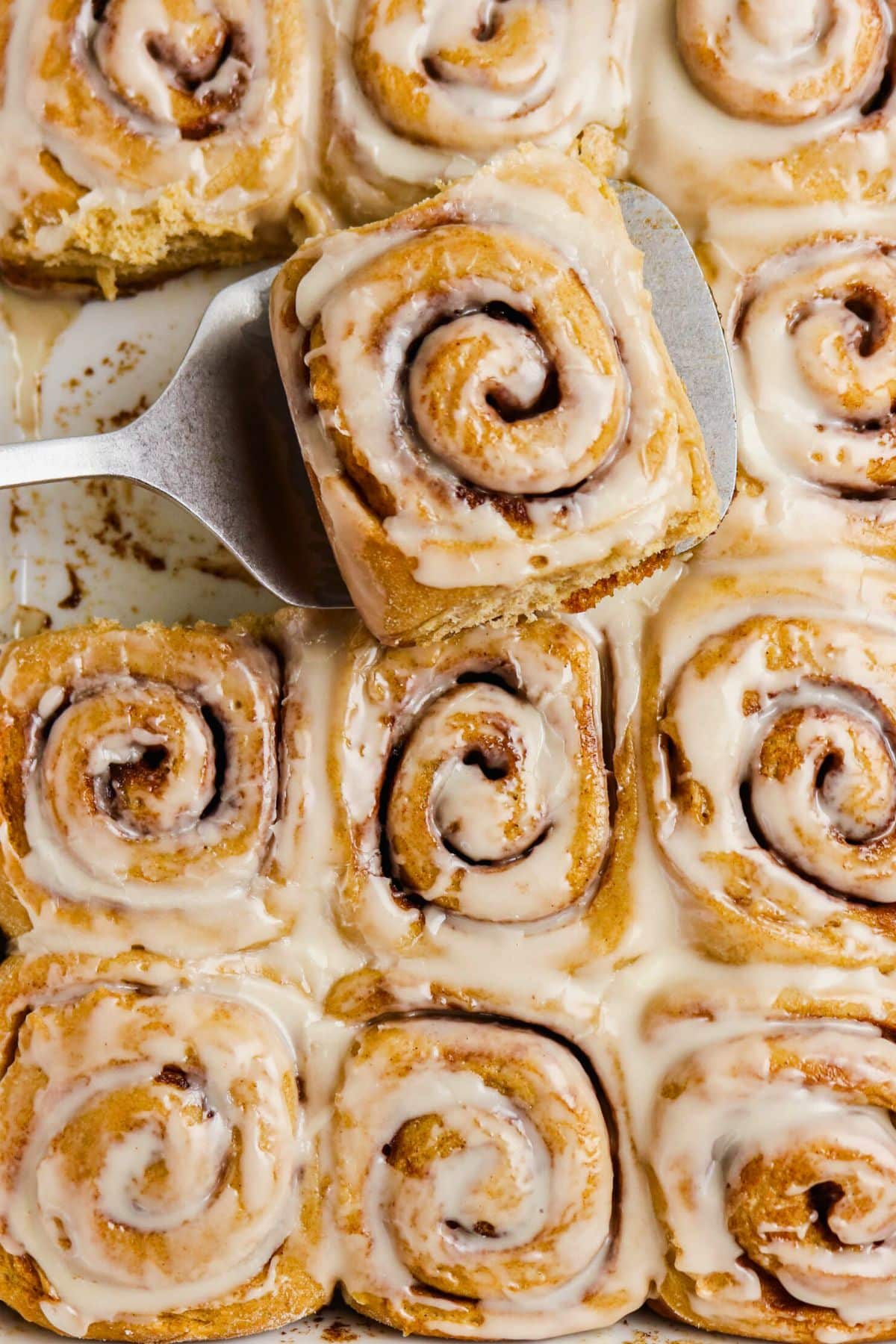 A spatula lifts a cinnamon roll from a tray of several glazed cinnamon rolls arranged closely together, all topped with a layer of white icing.