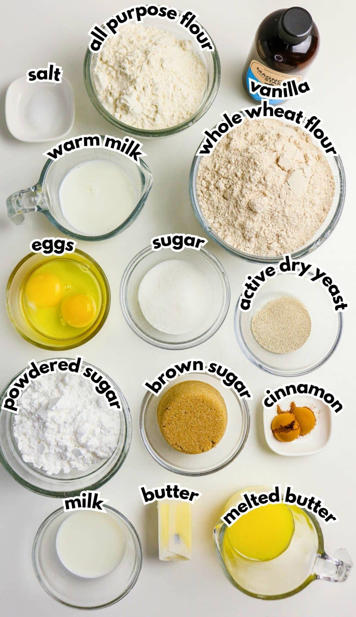 A top-down view of ingredients in bowls and measuring cups, labeled as all-purpose flour, whole wheat flour, vanilla, salt, warm milk, eggs, sugar, active dry yeast, powdered sugar, brown sugar, cinnamon, milk, butter, and melted butter.