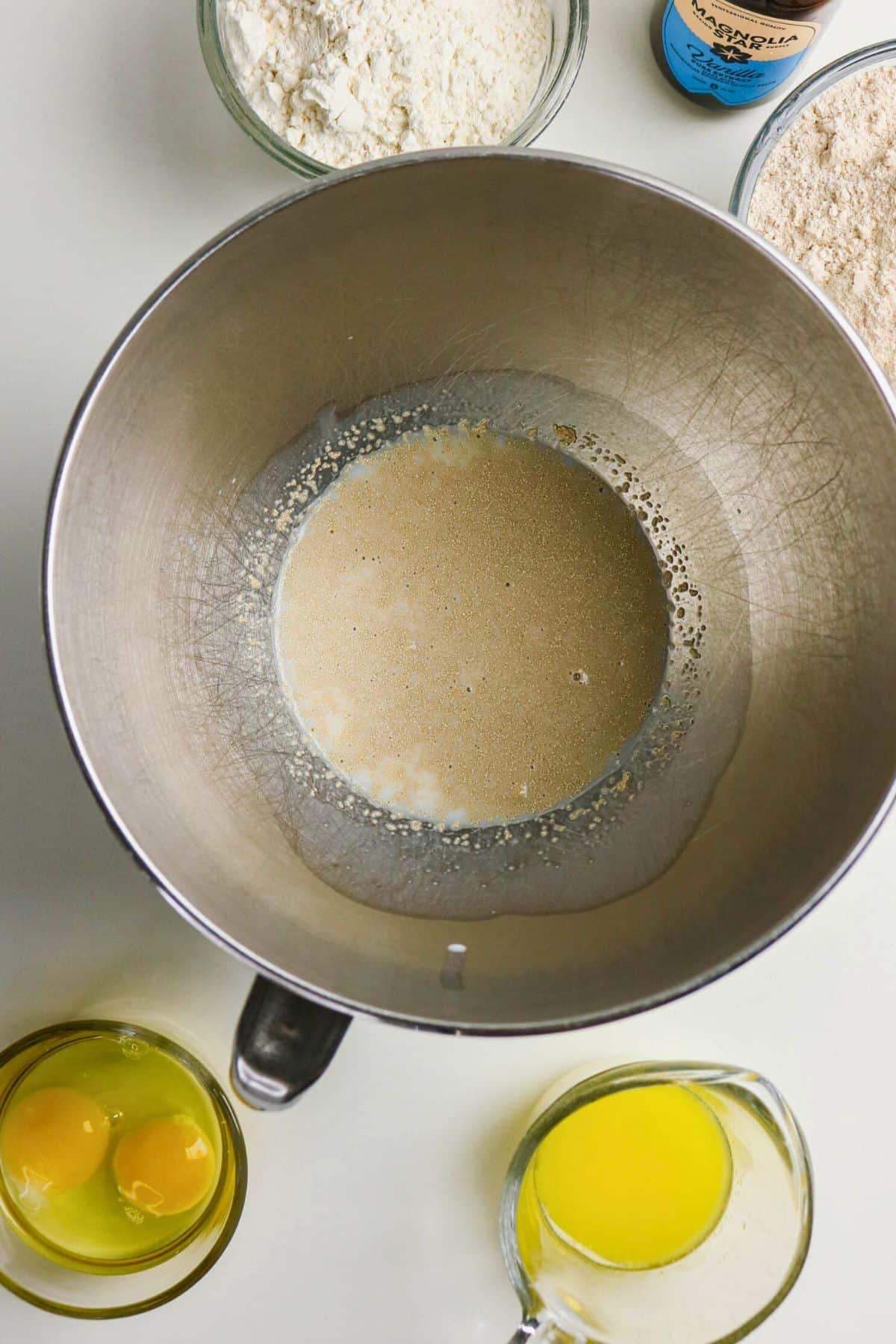 A mixing bowl with activated yeast sits on a counter, surrounded by bowls of flour, whole wheat flour, two cracked eggs, and a measuring cup of melted butter.