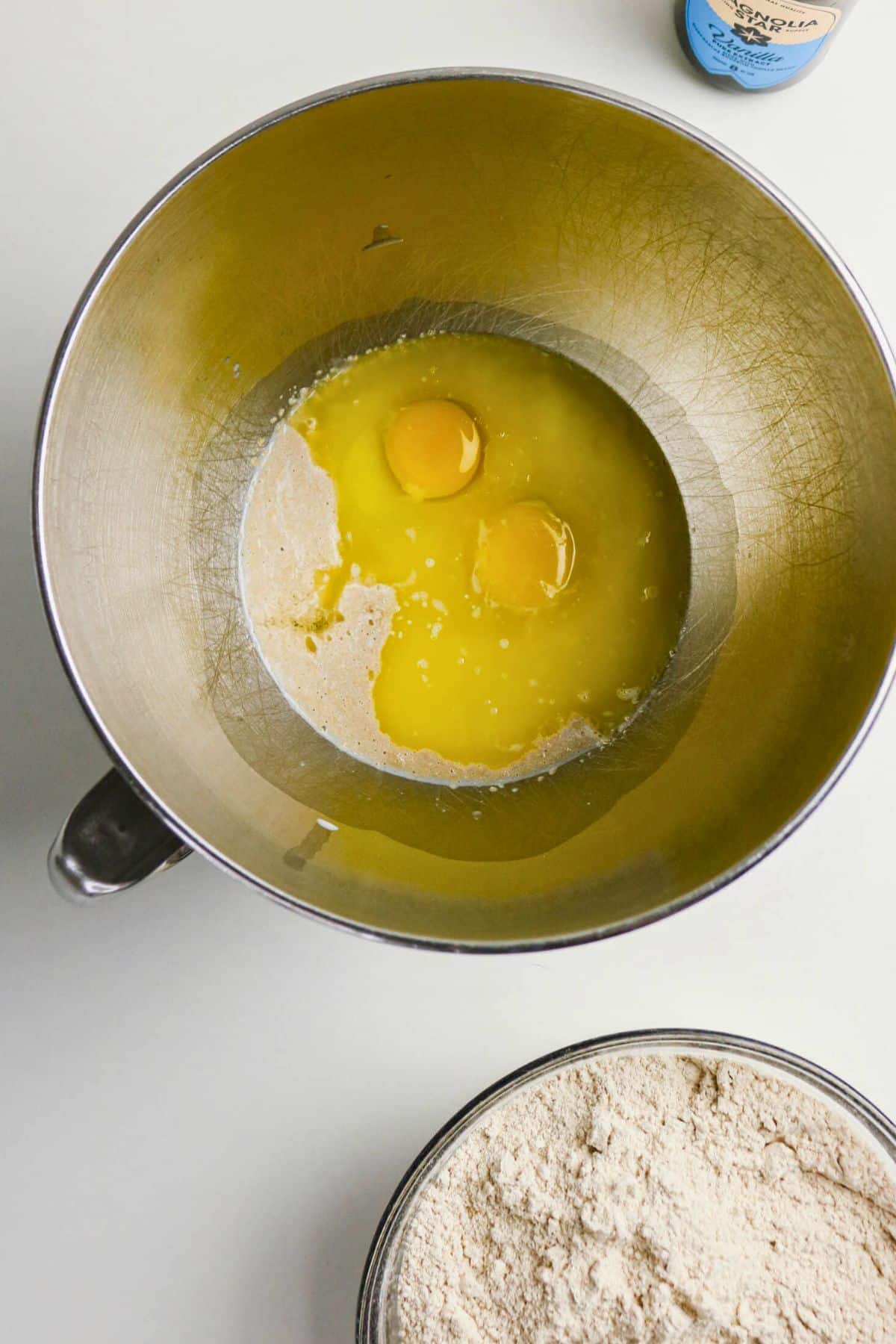A mixing bowl with eggs, melted butter, and sugar, next to another bowl filled with flour on a white surface.