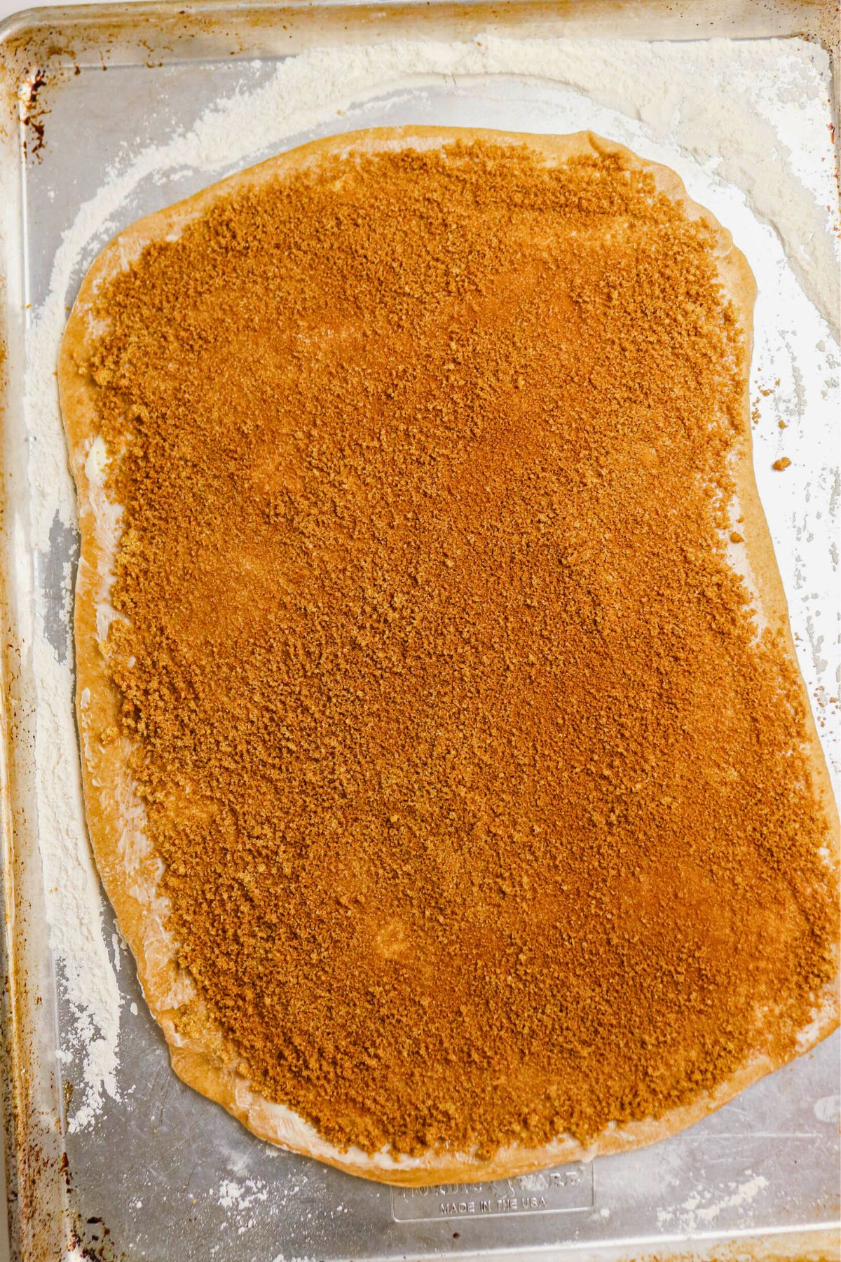 A sheet of rolled-out dough on a metal baking tray is topped evenly with a layer of brown sugar and cinnamon mixture, covering most of the surface.