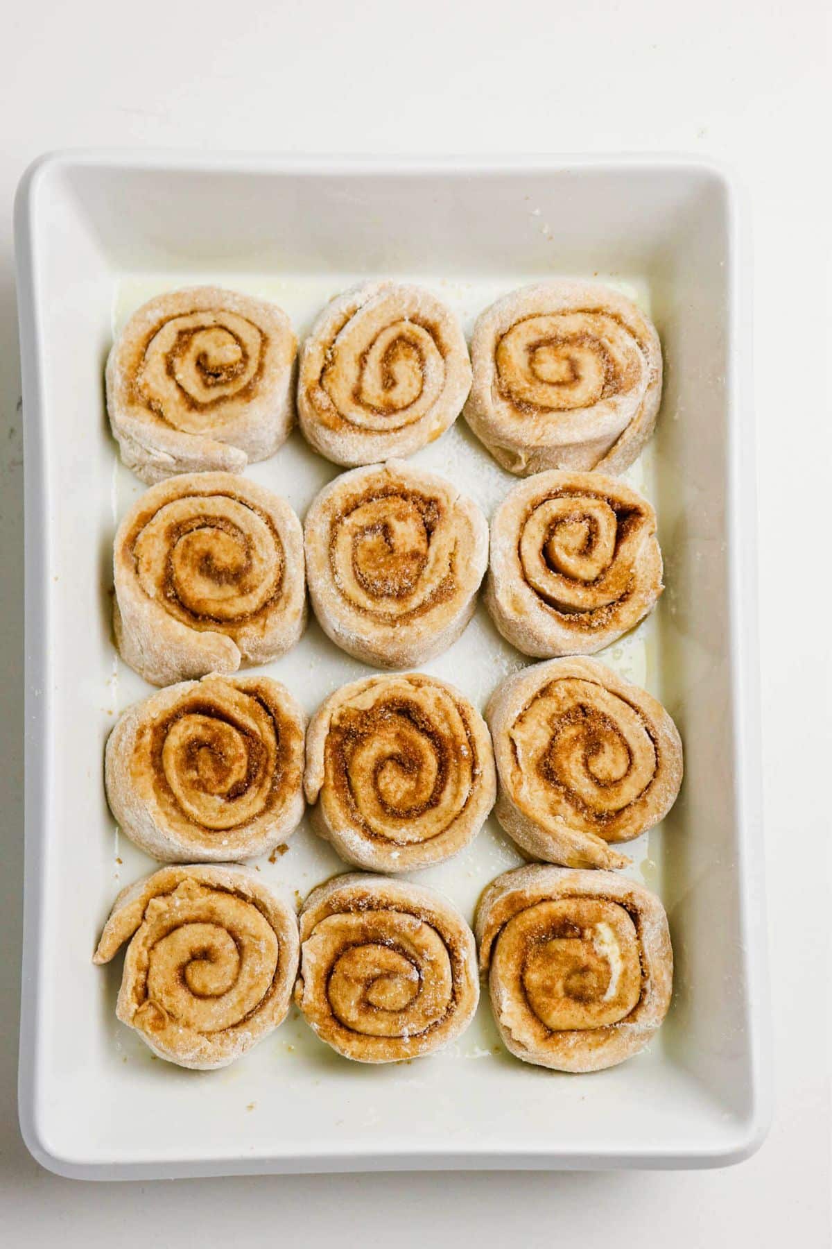 A rectangular white baking dish holds twelve unbaked cinnamon rolls, arranged in a 3 by 4 grid on a lightly floured surface. The rolls are swirled and covered with cinnamon sugar filling.