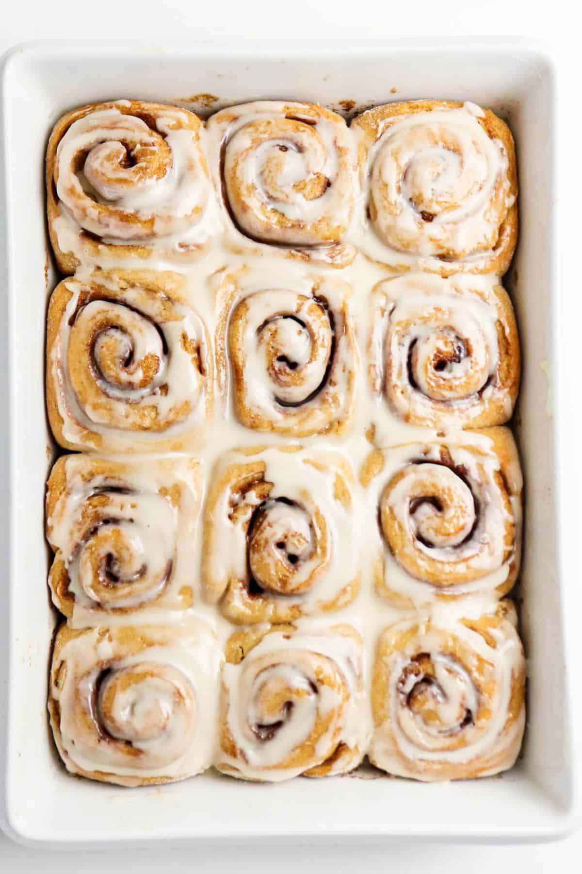 A rectangular baking dish filled with twelve iced cinnamon rolls, arranged in a 3 by 4 grid, with visible swirls and a layer of white icing on top.
