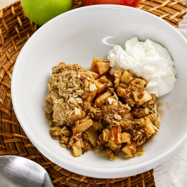 Apple crisp in a white bowl ready to enjoy with whipped cream on the side.