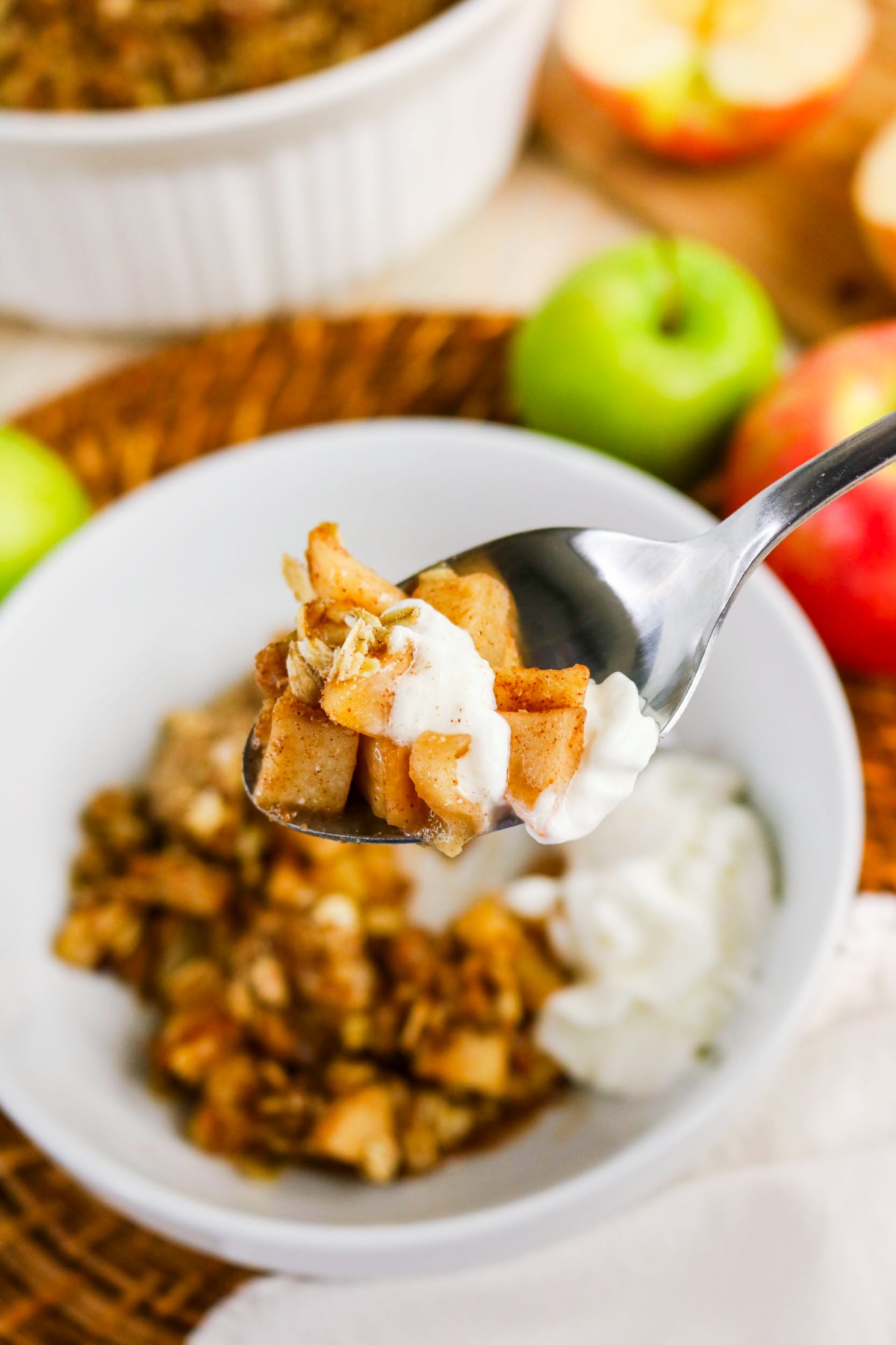 A spoonful of apple dessert with whipped cream over a white bowl of the same dessert.