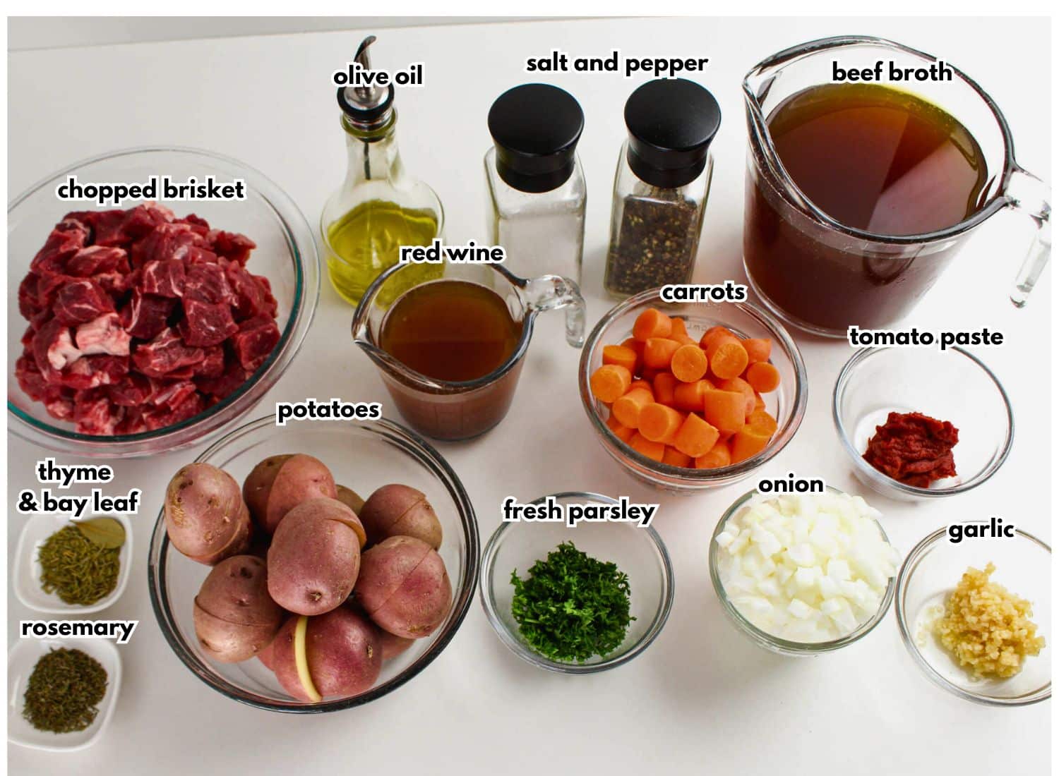 Ingredients for beef brisket stew arranged on countertop including brisket, potatoes, carrots, broth, and herbs.