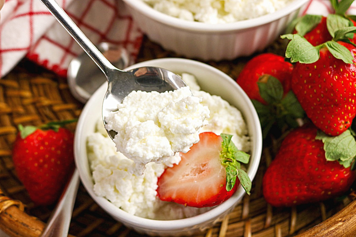 A serving of homemade cottage cheese in a white dish with a spoon and slice of strawberry on the side.