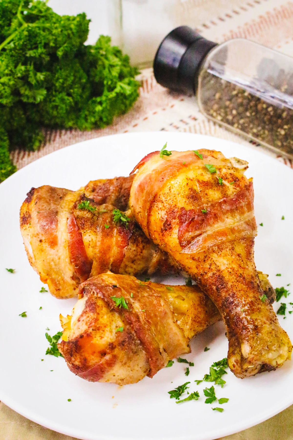 Three chicken drumsticks wrapped with bacon on a wite serving plate.