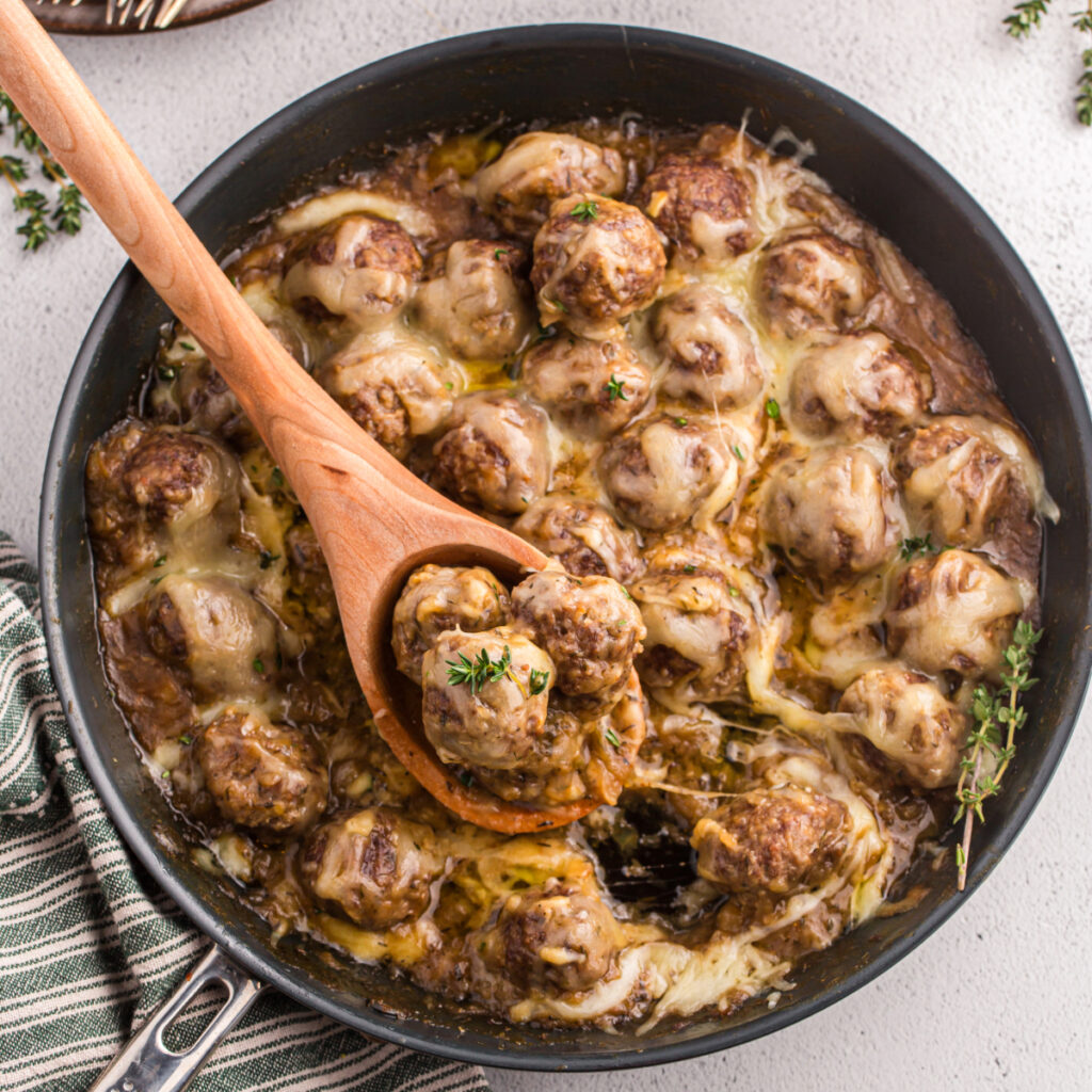 A wooden spoon scooping out french onion meatballs from a skillet.