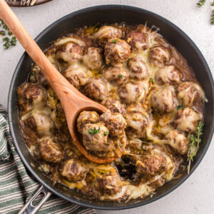 A wooden spoon scooping out french onion meatballs from a skillet.
