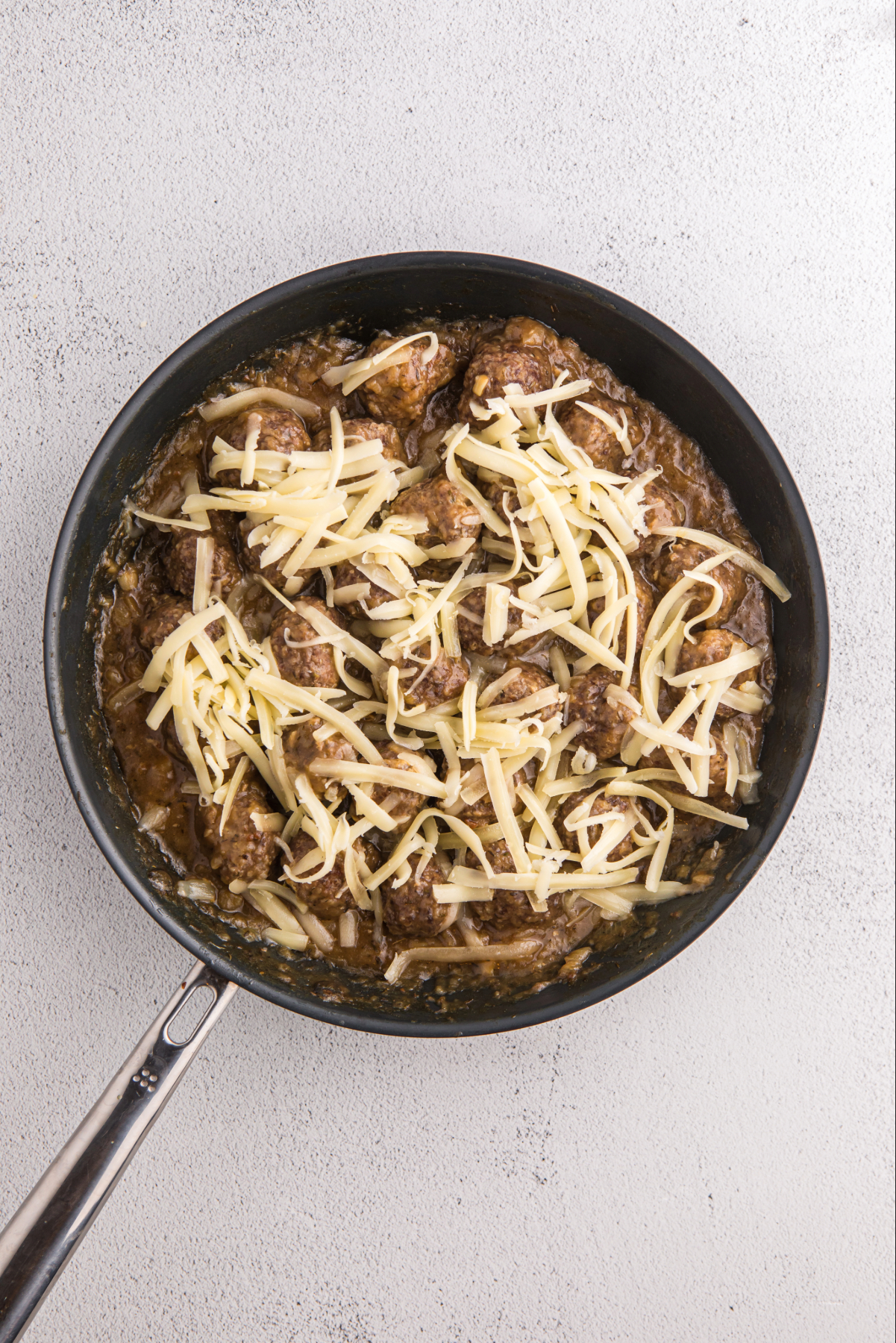 Shredded cheese over meatballs in a skillet.
