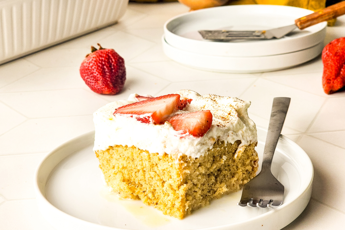 A square slice of tres leches cake topped with whipped topping and sliced strawberries on a white plate.