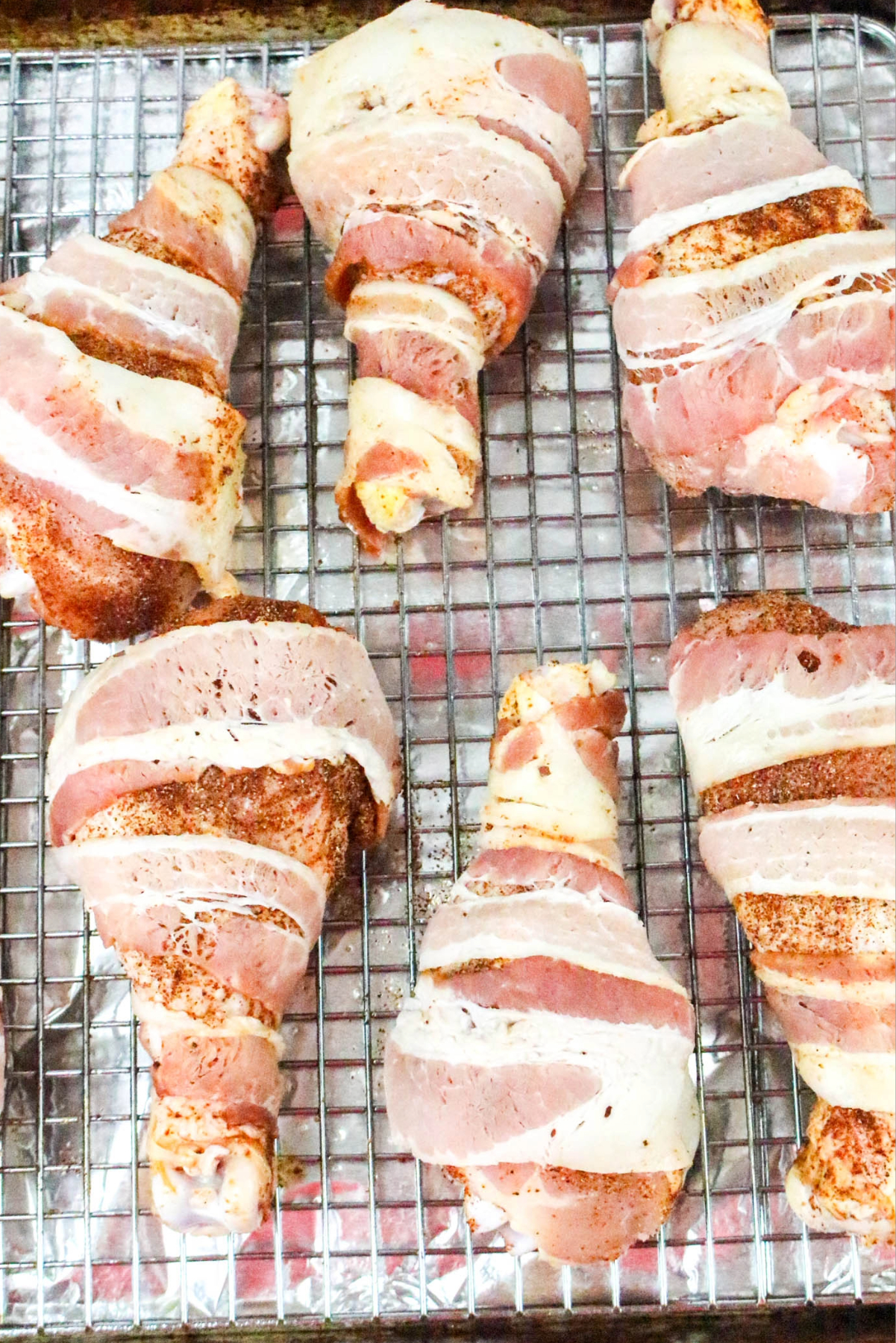 chicken drumsticks wrapped with bacon on a baking sheet before cooking.