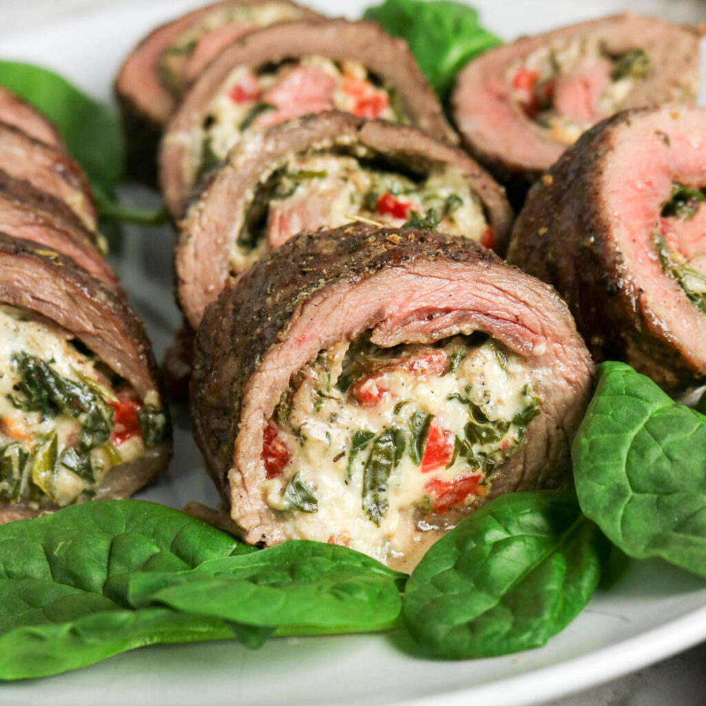 Stuffed flank steak on a white platter topped with fresh spinach leaves.