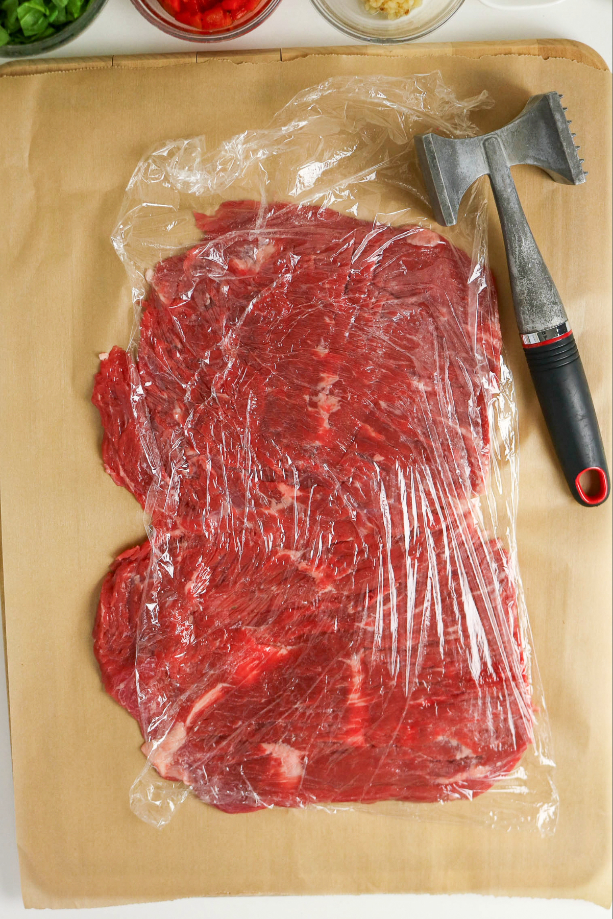 Flank steak on a parchment lined cutting board topped with plastic wrap.
