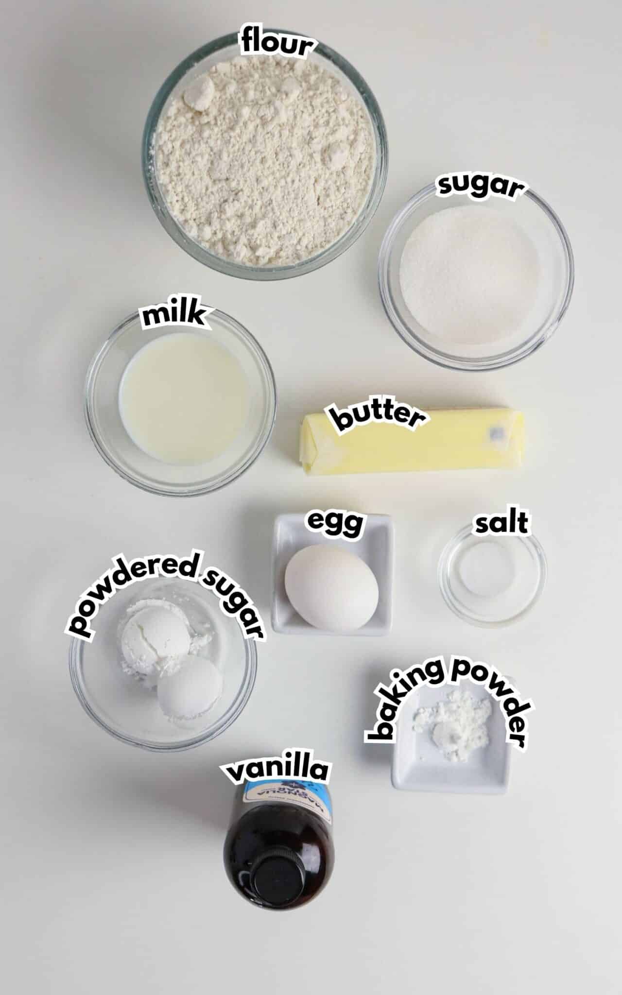 Bowls of flour, sugar, milk, butter, egg, salt, powdered sugar, baking powder and vanilla on a table.
