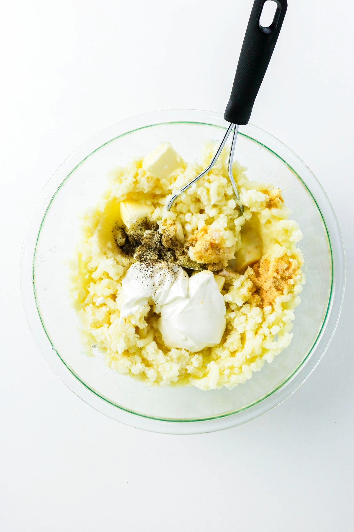 Adding sour cream and spices to mashed potatoes in a clear mixing bowl.