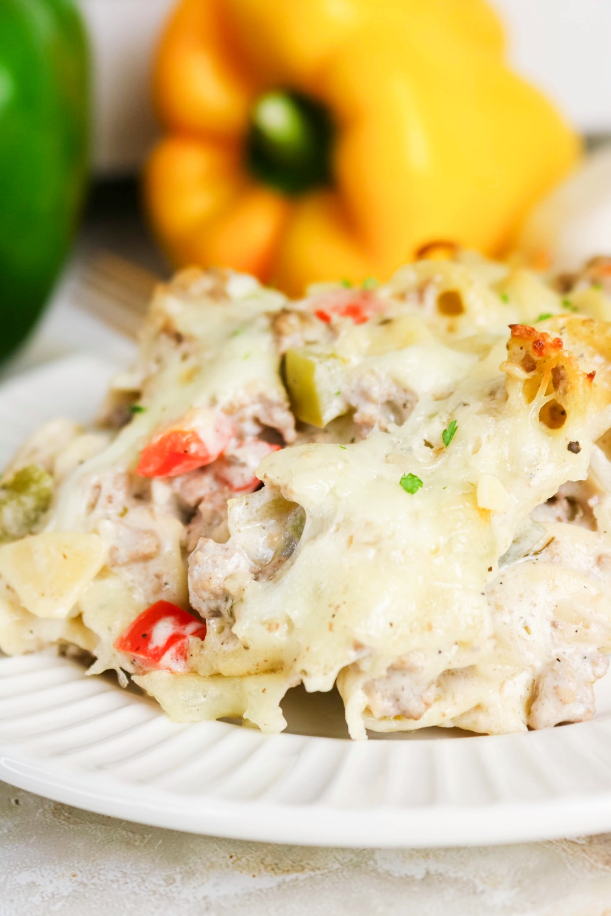A white dinner plate with a serving of philly cheesesteak casserole with yellow and green bell peppers in the back.