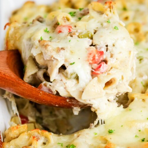 A wooden spoon scooping up a serving of philly cheesesteak casserole from a white baking dish.