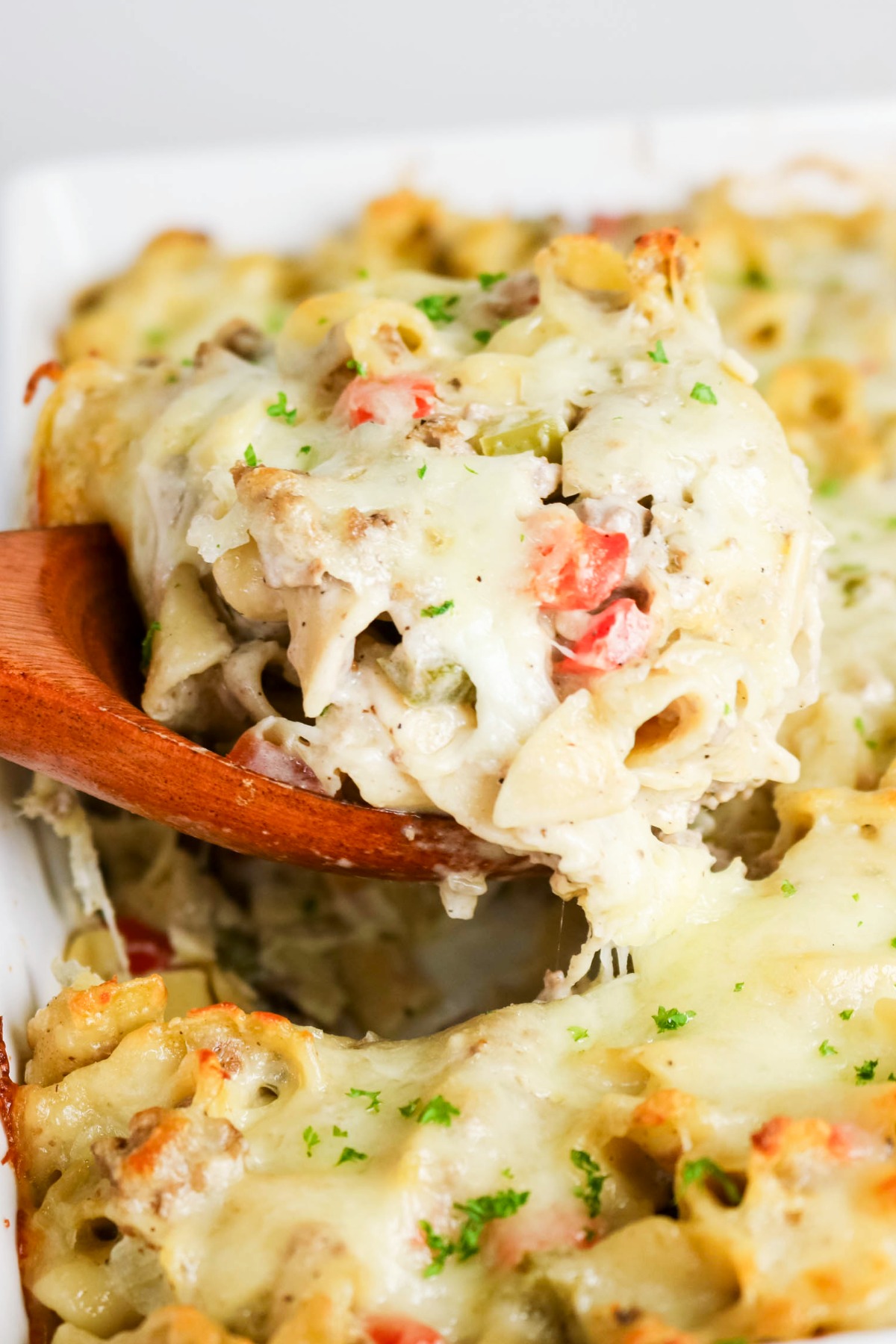 A wooden spoon scooping up a serving of philly cheesesteak casserole from a white baking dish.