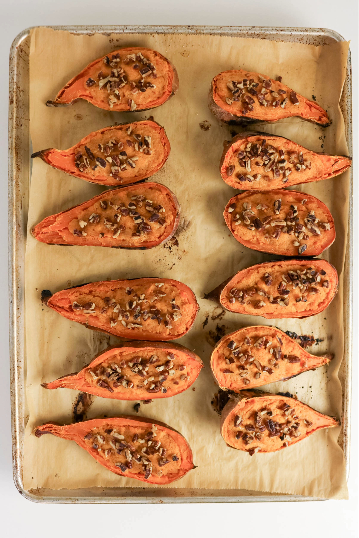 Sliced baked sweet potatoes topped with pecans on a parchment lined baking sheet.