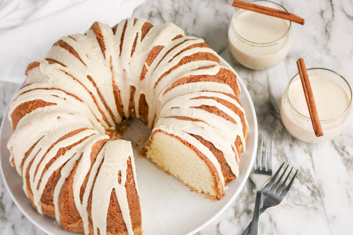 Two cups of eggnog topped with cinnamon sticks on the side of a pound cake.